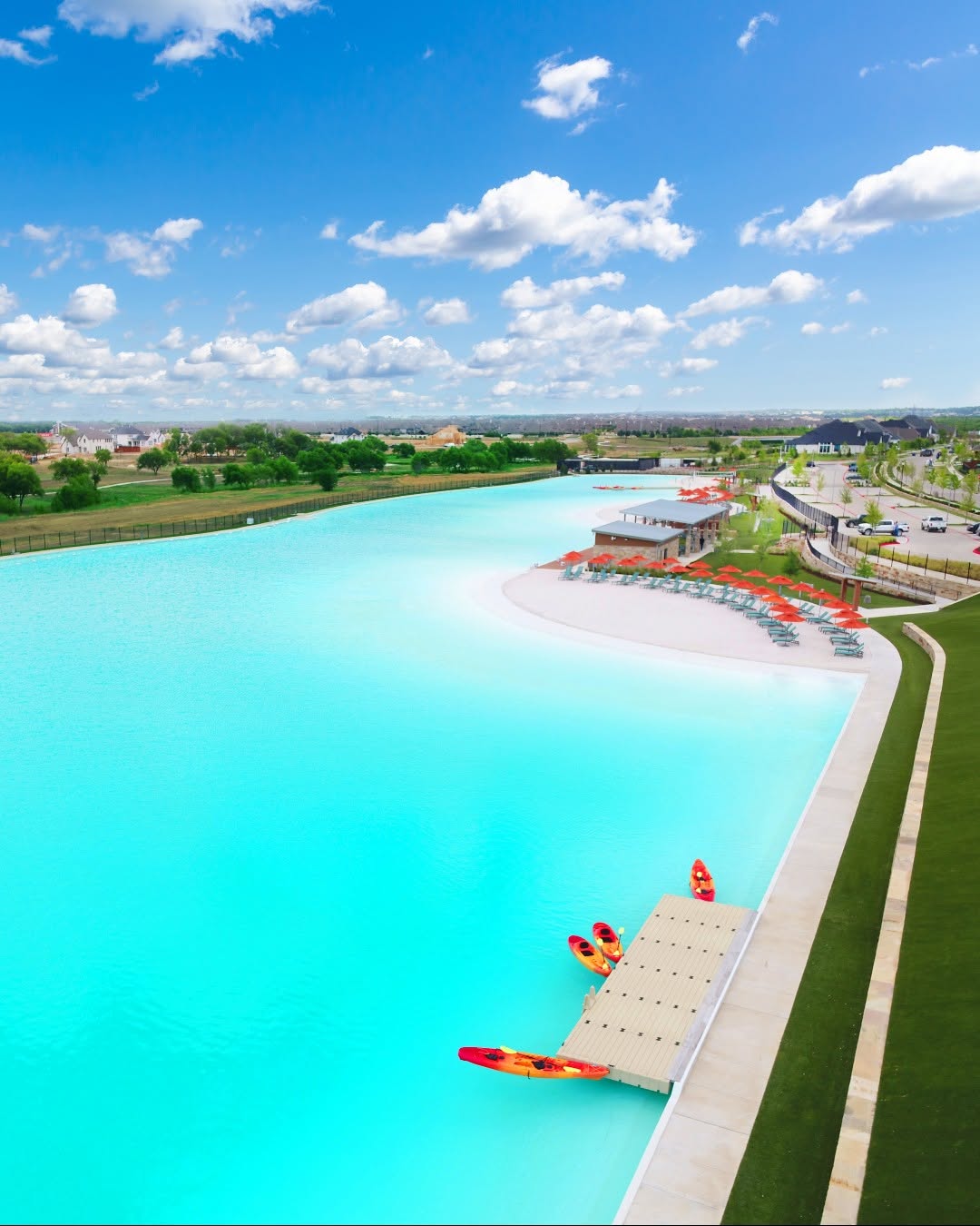 Aerial view of the bright turquoise Windsong Ranch Crystal Lagoon with kayaks, sandy beach, and lounge seating under a blue sky